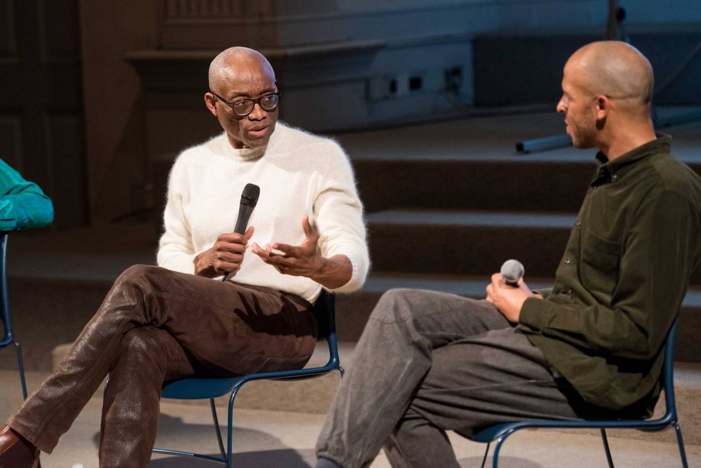 Bill T. Jones in glasses, a white sweater and brown pants speaks to Will Rawls